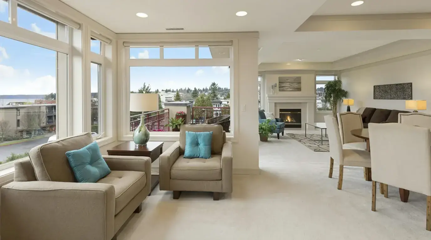 A bright living room with pale sofas, a fireplace and a corner window over a coastal town