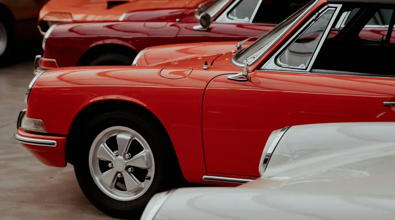 A row of classic cars parked nose to tail, a red coupe with chrome trim and a five-spoke alloy wheel closest to the camera.