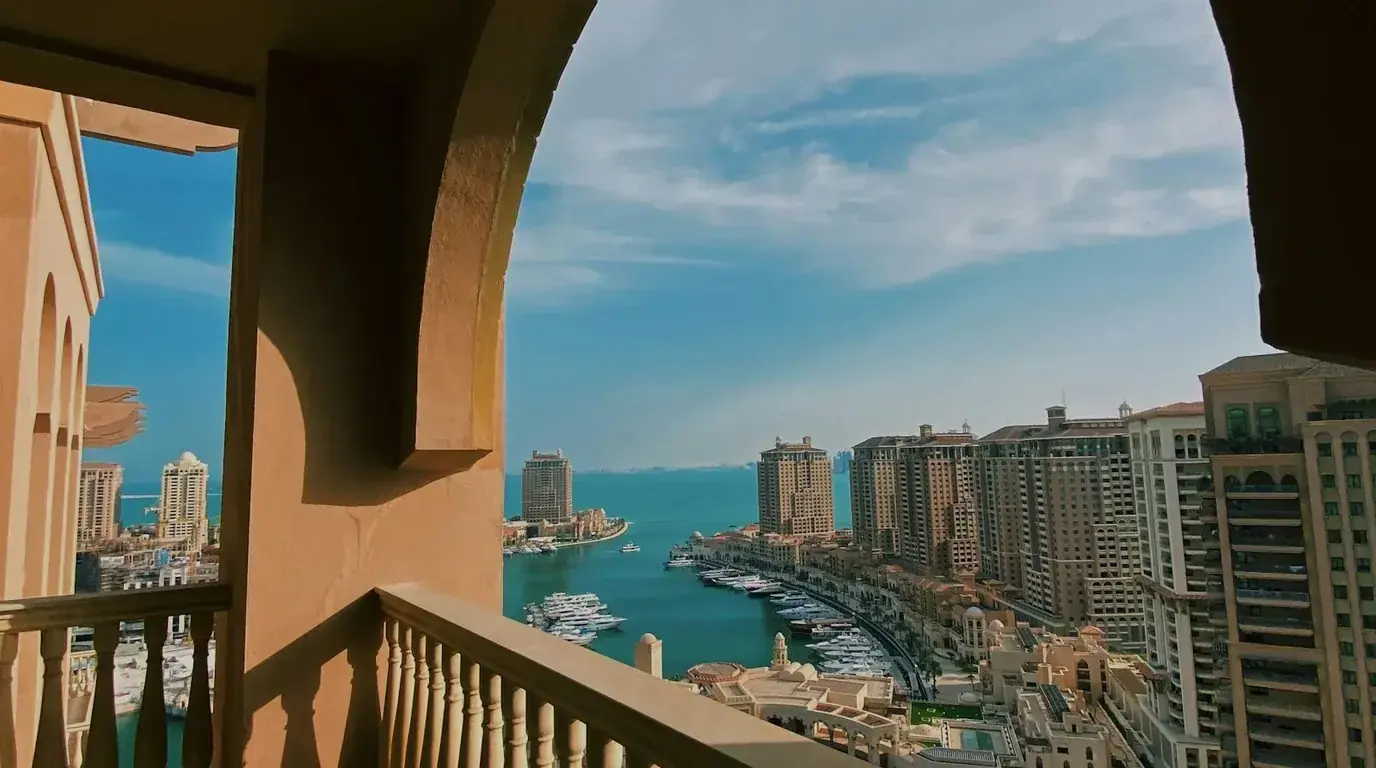 A view through a stone archway over a marina of moored yachts, ringed by tall sand-coloured apartment towers.