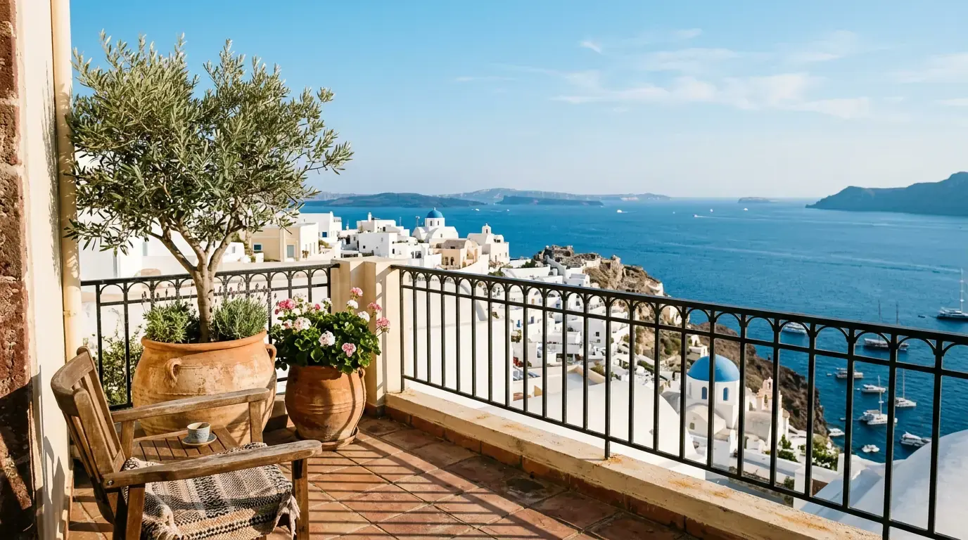 A sunlit terracotta-tiled balcony with an olive tree in a pot, rooftops and open sea beyond