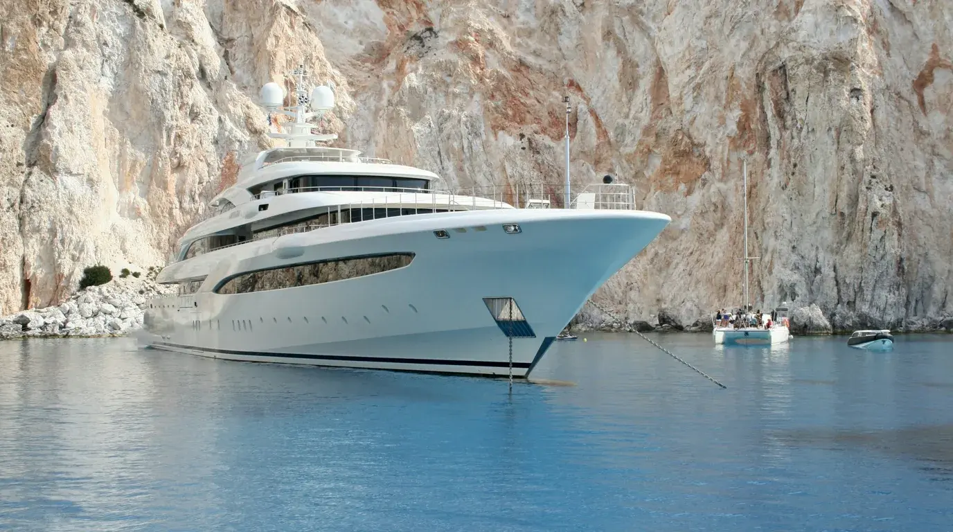 A large white superyacht at anchor in clear blue water beneath a pale limestone cliff.