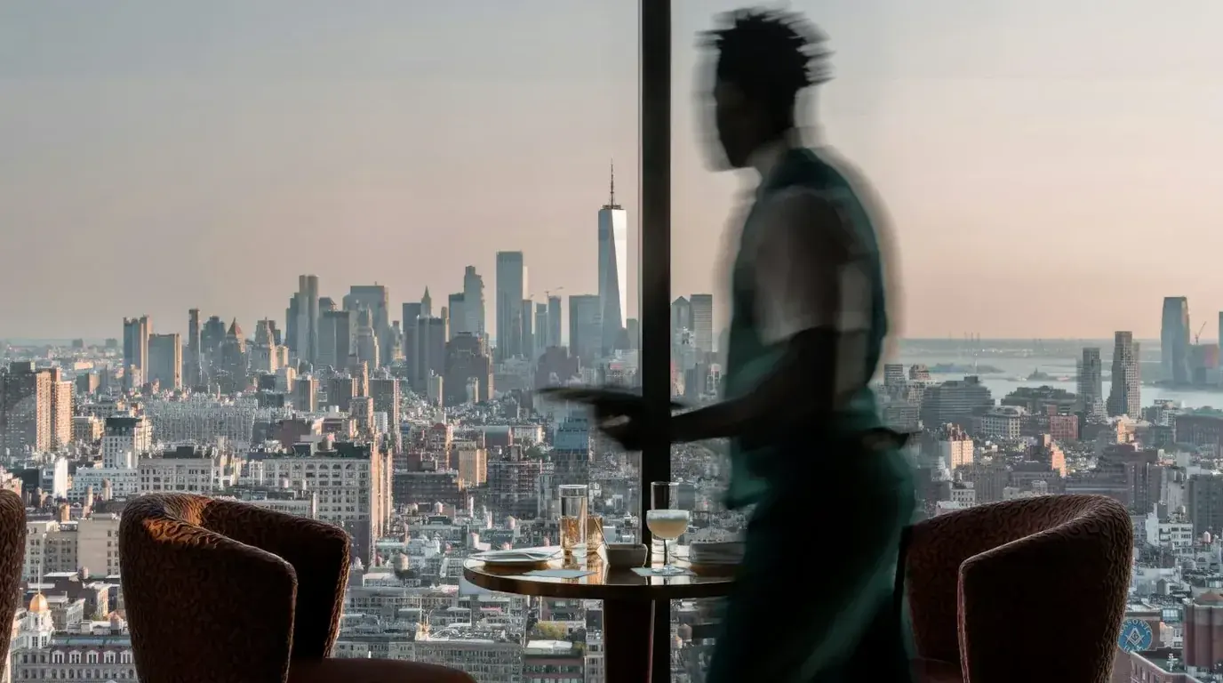 A man carrying a tray past a window overlooking the Manhattan skyline at dusk