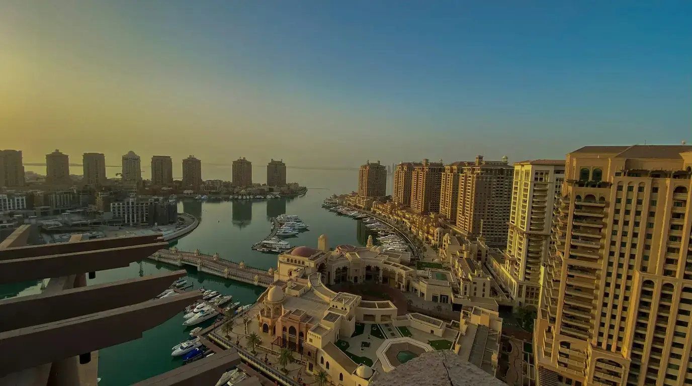 A marina seen from a high balcony at sunset, yachts moored along a curved quay below tall sand-coloured apartment towers.